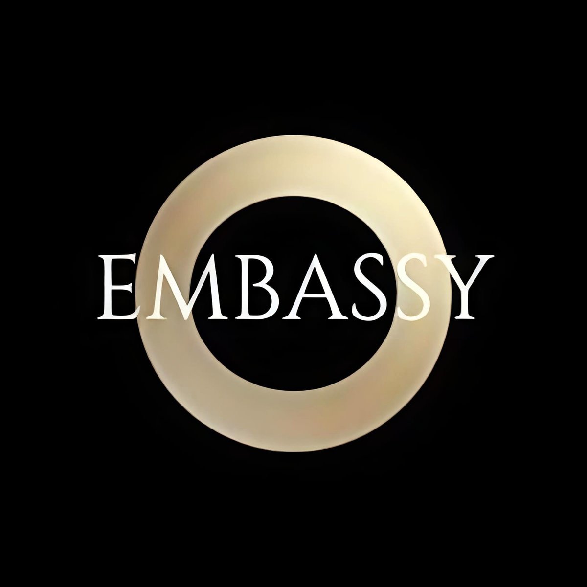 EMBASSY logo featuring white text with a tan circular ring design on black background